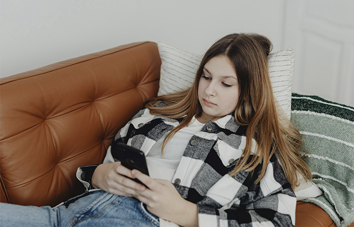 Teenage girl on the sofa scrolling on her phone, represents how to much social media time can be a risk factor for teen depression.
