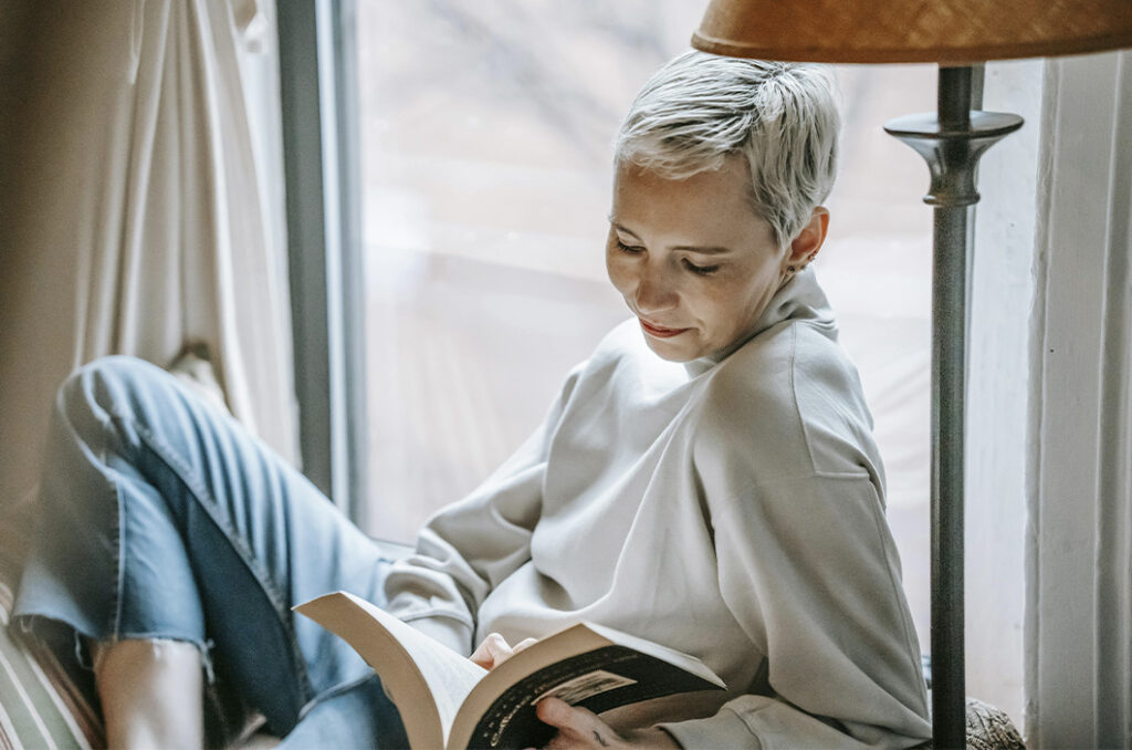 Woman reading a book by the window, represents our selection of best grief books for adults to help you navigate loss.