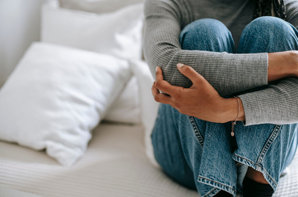 Woman siting on a bed, embracing her knees, represents the signs of attachment trauma in adults.