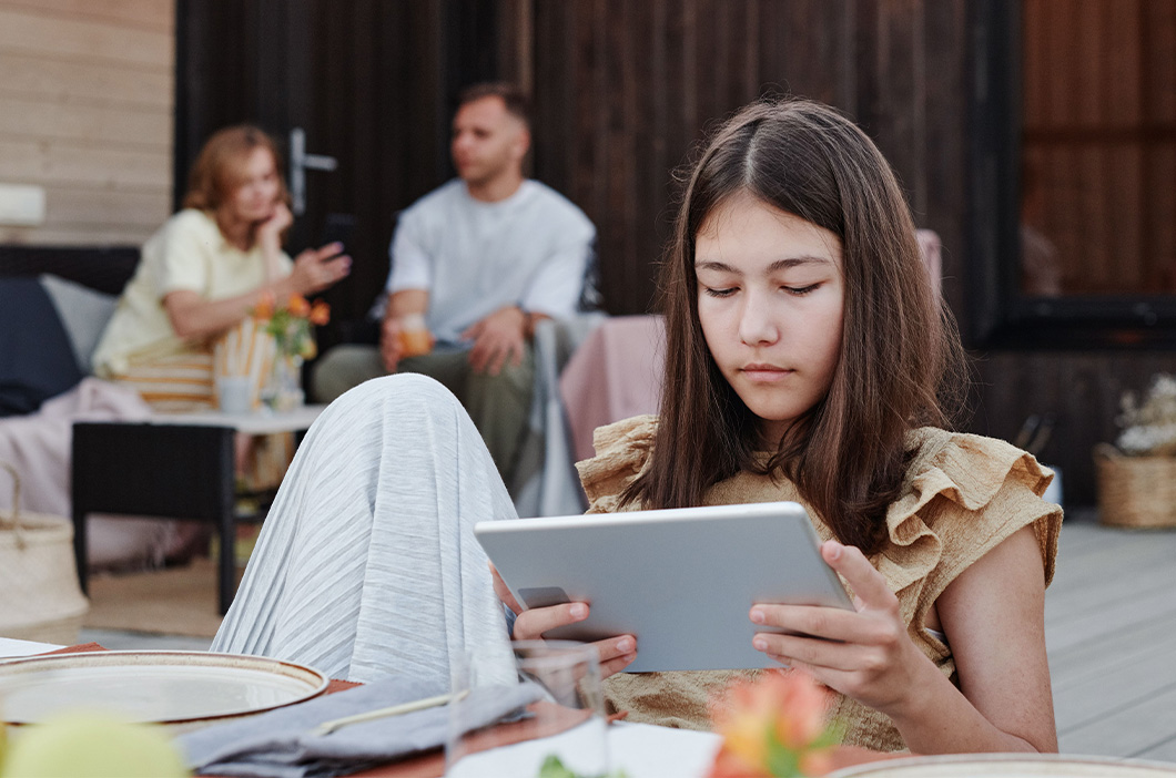 Teen girl on her tablet with her parent on the background talking, represents how to help a teenager with social anxiety.
