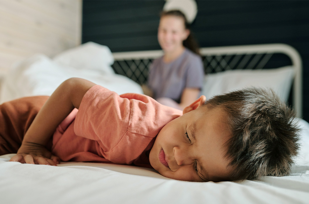 Young boy lying in bed looking upset with a parent in the background, represents the hidden trauma of emotionally unavailable parents.